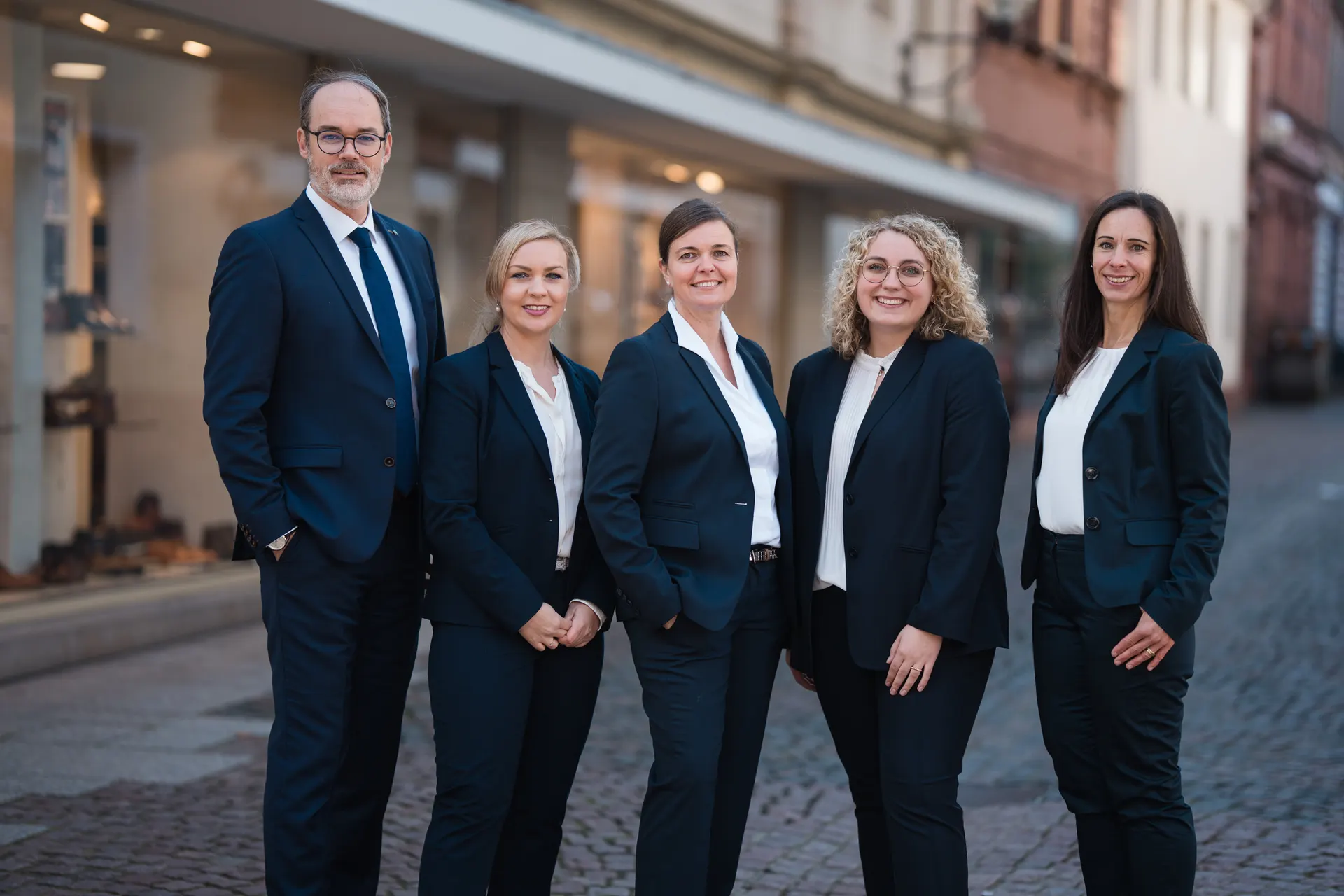 Teamfoto von fünf Mitarbeitenden in dunklen Anzügen und Blazern in einer Altstadtgasse